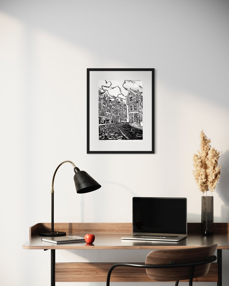 May include: Black and white line art print of a city street with buildings on either side. The print is framed in a black frame and hanging on a white wall.