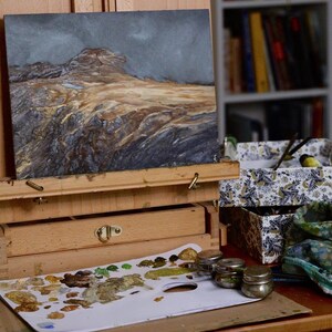 May include: A wooden easel with a painting of a landscape on it. The painting is of a rocky, mountainous area with a blue sky and some clouds. There are also some paint tubes and a palette with paint on it.