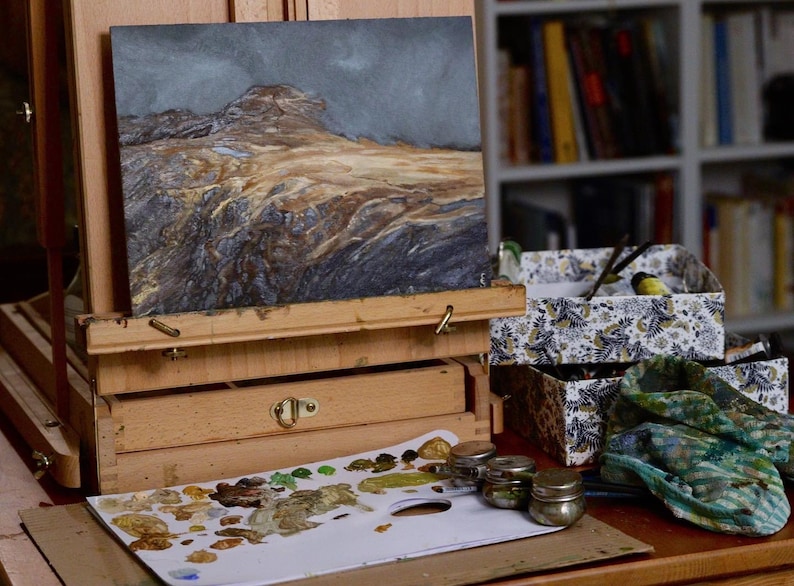 May include: A wooden easel with a painting of a landscape on it. The painting is of a rocky, mountainous area with a blue sky and some clouds. There are also some paint tubes and a palette with paint on it.