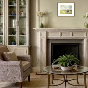 May include: A living room with a white fireplace, a glass-front cabinet, a wicker armchair, and a glass coffee table with a green plant. A framed painting of a landscape with a blue sky and a white house hangs above the fireplace.