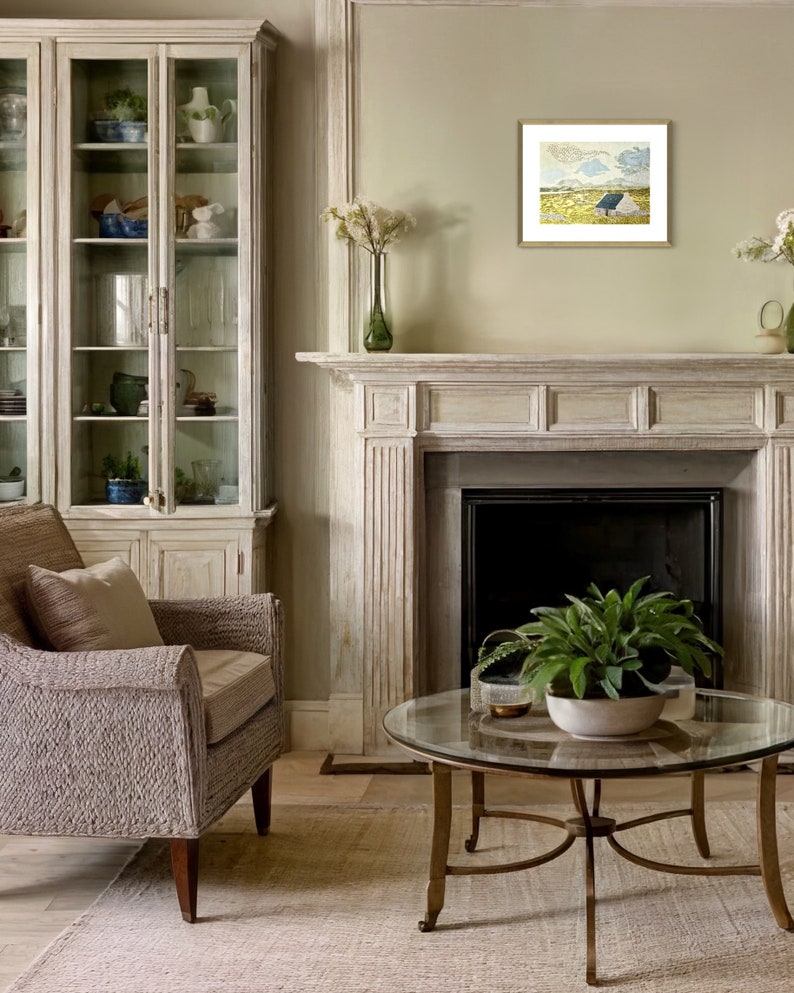 May include: A living room with a white fireplace, a glass-front cabinet, a wicker armchair, and a glass coffee table with a green plant. A framed painting of a landscape with a blue sky and a white house hangs above the fireplace.