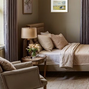May include: A beige bedroom with a white bed, a beige armchair, a side table with a lamp, and a framed painting of a landscape with a small house on a hill.