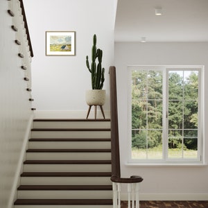 May include: A white staircase with dark wood steps and a white railing. A potted cactus plant sits on a wooden stand in the hallway. A framed landscape painting hangs on the wall above the stand. A window with white trim looks out onto a green lawn and trees.