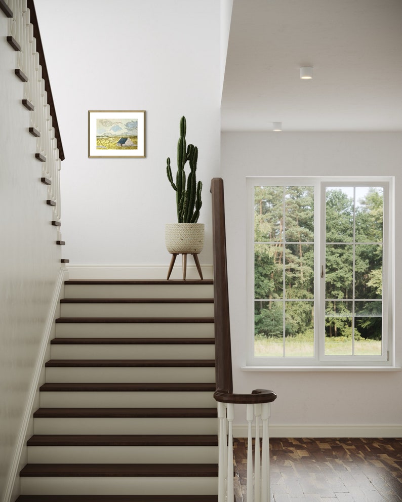May include: A white staircase with dark wood steps and a white railing. A potted cactus plant sits on a wooden stand in the hallway. A framed landscape painting hangs on the wall above the stand. A window with white trim looks out onto a green lawn and trees.