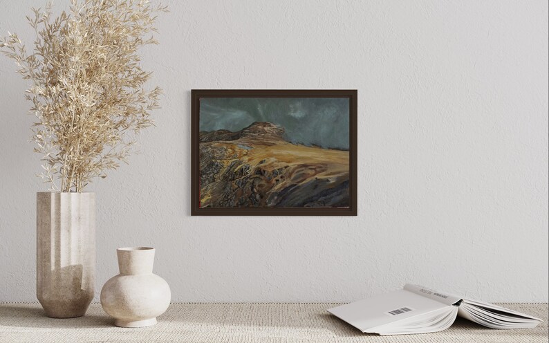 May include: A framed painting depicting a mountainous landscape with a cloudy sky. The mountain is brown and the sky is a pale blue with white clouds. The painting is in a dark brown frame.