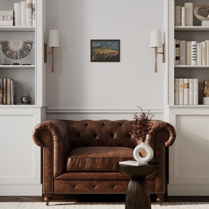 May include: A brown leather armchair with buttoned upholstery sits in a room with white built-in bookshelves, gold sconces, and a small painting on the wall.