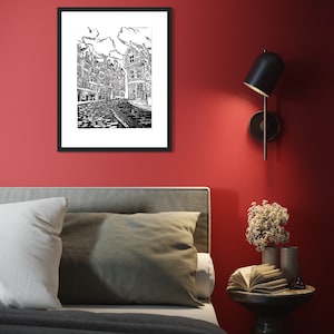 May include: Black and white line art print of a city street with buildings and cobblestone street. The print is framed in a black frame and hanging on a red wall.