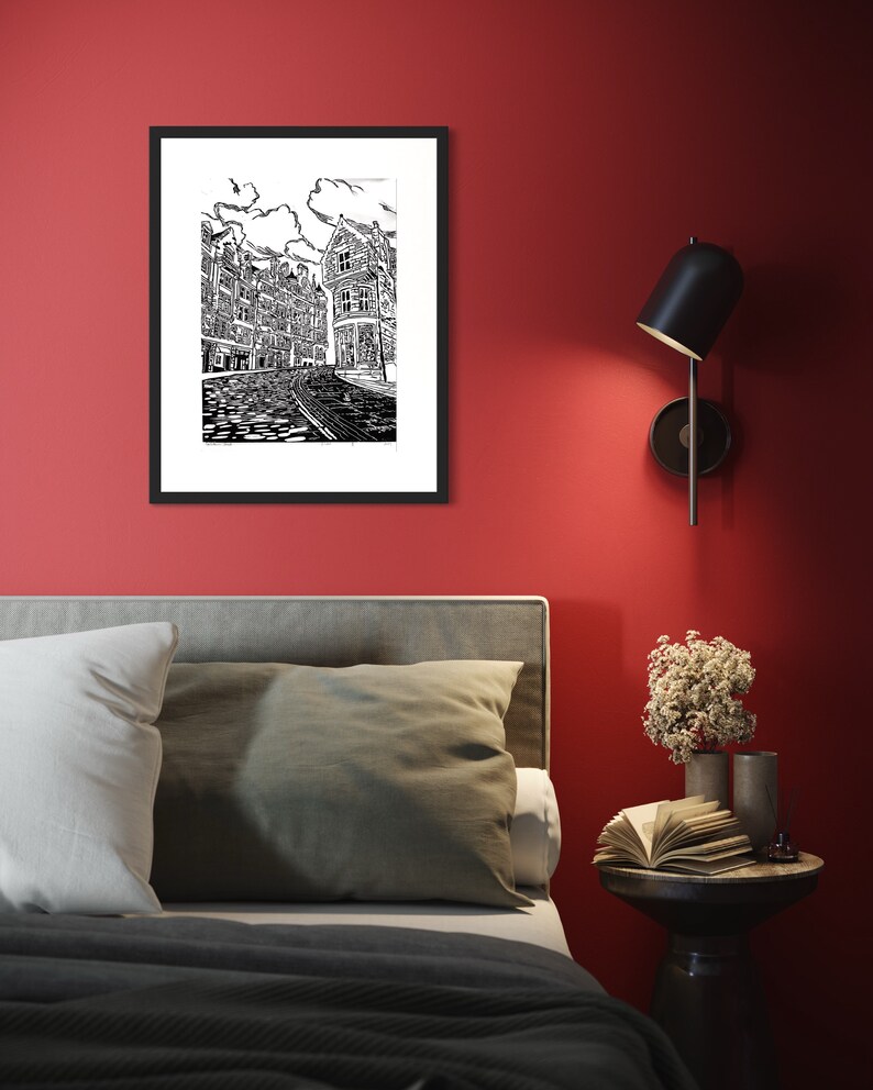 May include: Black and white line art print of a city street with buildings and cobblestone street. The print is framed in a black frame and hanging on a red wall.