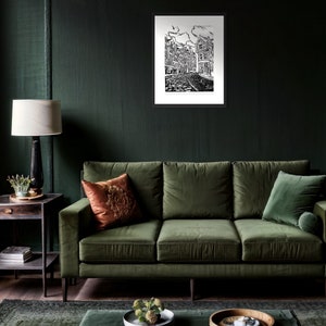 May include: A black and white illustration of a city street scene with buildings and cobblestone streets. The illustration is framed and hanging on a dark green wall above a green velvet sofa.