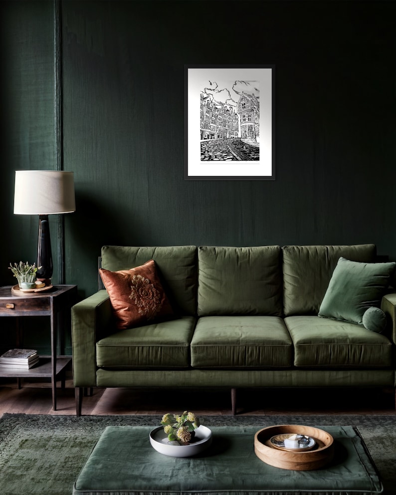 May include: A black and white illustration of a city street scene with buildings and cobblestone streets. The illustration is framed and hanging on a dark green wall above a green velvet sofa.