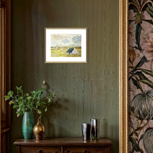 May include: A framed print of a watercolor painting depicting a small, white cottage with a blue roof in a field of yellow grass. The sky is a light blue with white clouds.