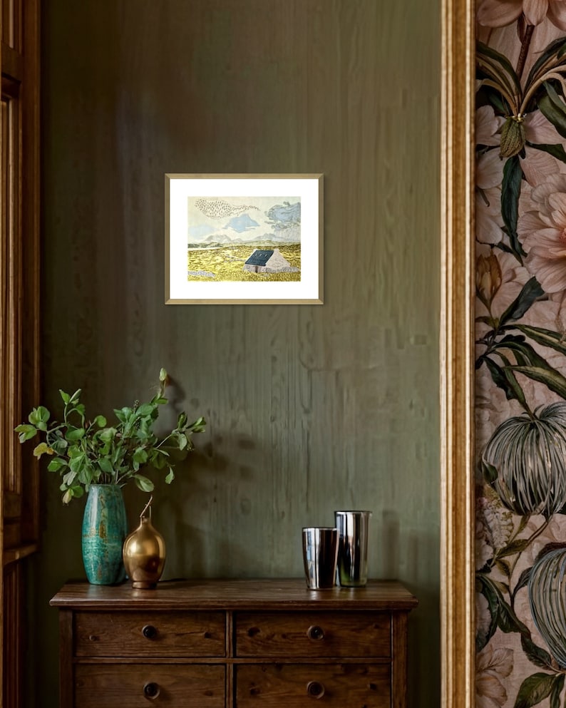 May include: A framed print of a watercolor painting depicting a small, white cottage with a blue roof in a field of yellow grass. The sky is a light blue with white clouds.