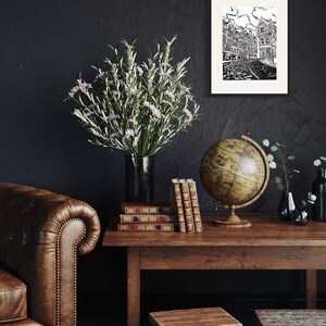 May include: A black and white illustration of a city street with buildings, a globe, and a stack of books on a wooden table. A vase with green and white flowers sits on the table. The background is a dark gray wall.
