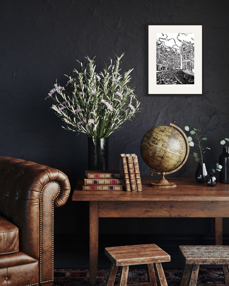 May include: A black and white illustration of a city street with buildings, a globe, and a stack of books on a wooden table. A vase with green and white flowers sits on the table. The background is a dark gray wall.