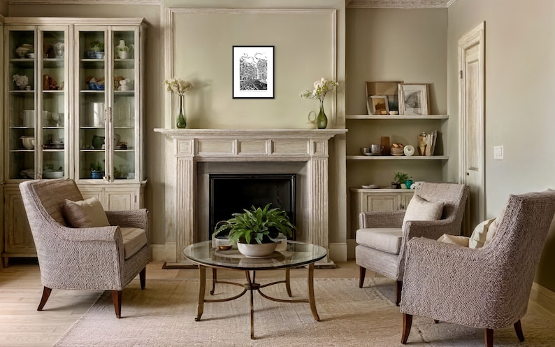 May include: A living room interior with two armchairs, a coffee table, a fireplace, and a glass-front cabinet. The walls are painted a light green color. The furniture is upholstered in a light gray fabric. The coffee table is made of glass and metal. The fireplace is made of white stone. The cabinet is made of wood and glass. A black and white framed print hangs above the fireplace.