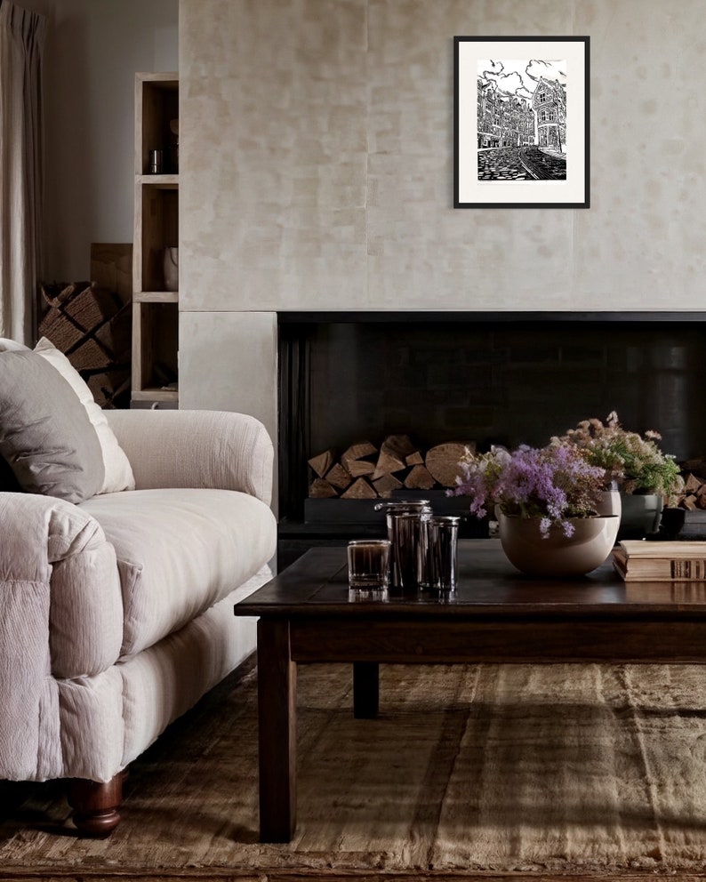 May include: A black and white illustration of a city street with buildings and cobblestone streets. The illustration is framed in a black frame and hanging on a wall above a fireplace.