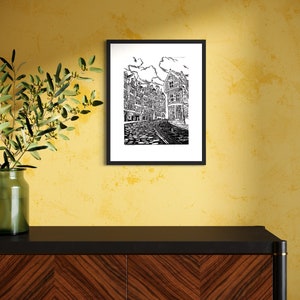 May include: Black and white line art print of a European city street with buildings and cobblestone street. The print is framed in a black frame and hanging on a yellow wall.