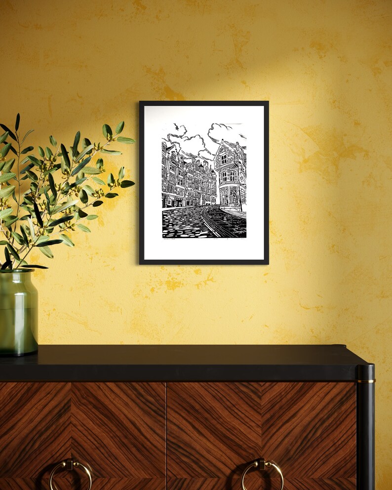 May include: Black and white line art print of a European city street with buildings and cobblestone street. The print is framed in a black frame and hanging on a yellow wall.