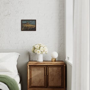 May include: A white bedroom with a bed, a bedside table, a lamp, and a framed landscape painting on the wall. The bedside table is made of wood and has two doors. The lamp is a white globe on a gold base. The painting is a small, framed landscape with a mountain in the background.