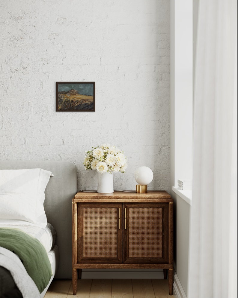 May include: A white bedroom with a bed, a bedside table, a lamp, and a framed landscape painting on the wall. The bedside table is made of wood and has two doors. The lamp is a white globe on a gold base. The painting is a small, framed landscape with a mountain in the background.