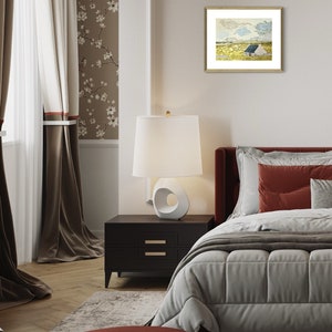 May include: A bedroom interior with a white lamp on a black nightstand, a bed with a white duvet and red pillows, and a framed print of a landscape with a small house on a hill.