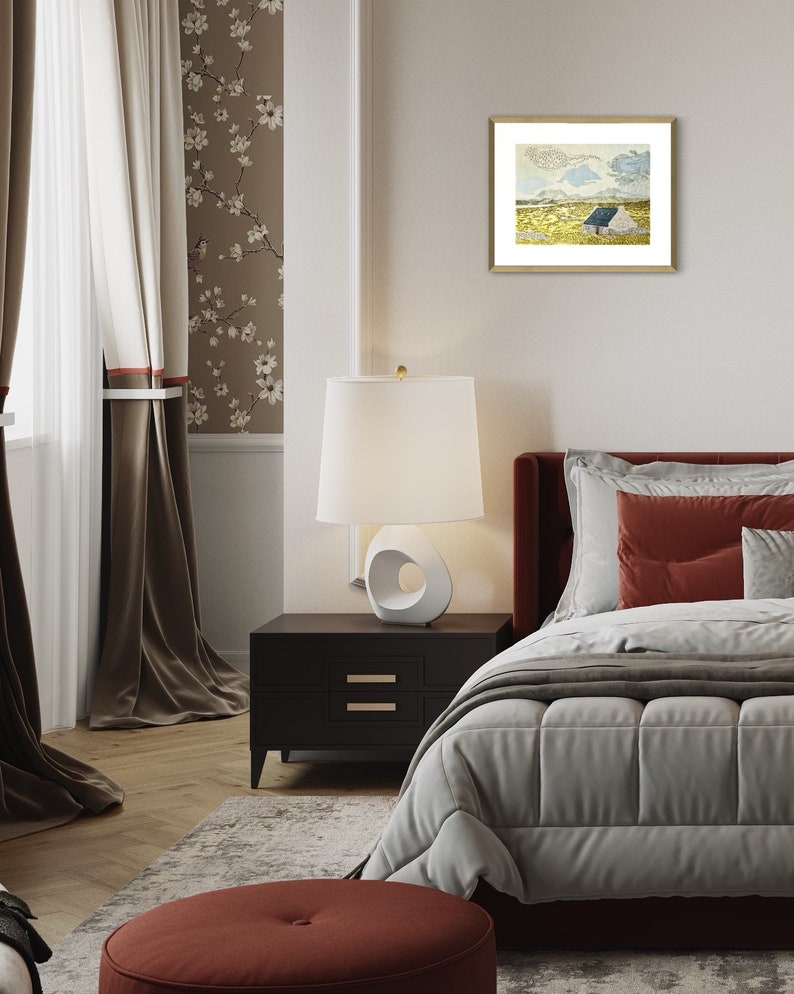 May include: A bedroom interior with a white lamp on a black nightstand, a bed with a white duvet and red pillows, and a framed print of a landscape with a small house on a hill.
