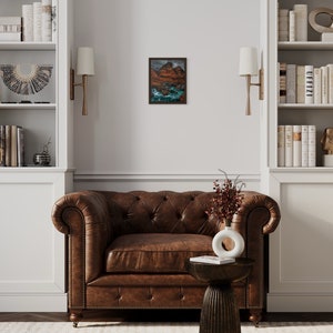 May include: A brown leather armchair with tufted upholstery sits in a room with white built-in bookshelves, a small side table, and a framed landscape painting.