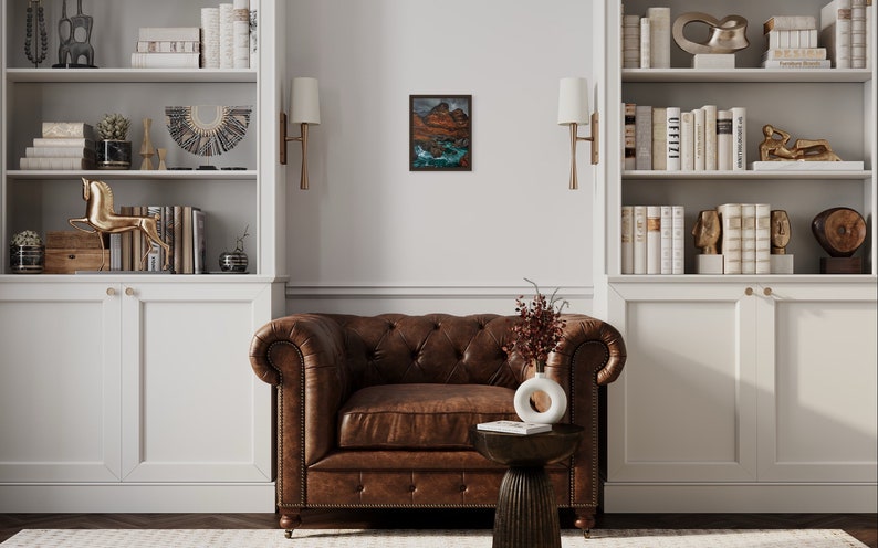 May include: A brown leather armchair with tufted upholstery sits in a room with white built-in bookshelves, a small side table, and a framed landscape painting.