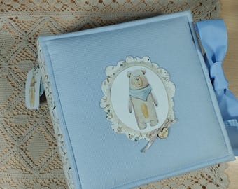May include: A light blue baby photo album with a decorative bear illustration on the cover. The album has a light blue ribbon closure and a decorative tag. The cover features a textured fabric.