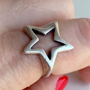 Könnte beinhalten: Ein silberner Ring mit einem sternförmigen Design. Der Stern ist in der Mitte offen, wodurch eine ausgeschnittene Form entsteht.