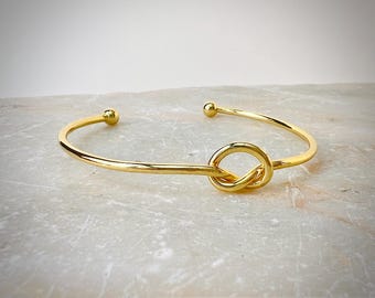 Peut inclure: Un bracelet jonc doré avec un motif de nœud au centre. Le bracelet a des extrémités arrondies avec de petits accents de boules. Le bijou est présenté sur une surface claire et texturée.