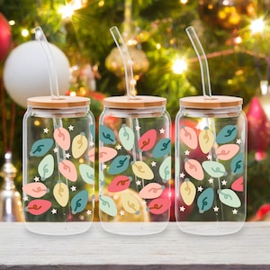 May include: Three clear glass drinking jars with bamboo lids and curved glass straws. Each jar is decorated with colorful Christmas light bulb designs in red, pink, yellow, teal, and green, with white star accents. The jars sit on a white surface.