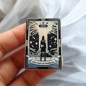 May include: A rectangular metal pin with a tarot card design. The pin has a black border with the words "XIII" and "DEATH" at the top. The image shows a figure standing in front of a starry background, with the text "THE COST OF FREEDOM" at the bottom.