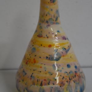 Peut inclure: Un vase en céramique fait à la main avec un motif tacheté. Le vase a une base jaune qui se fond dans une couleur crème, avec des mouchetures bleues, orange et violettes. Le vase mesure environ 20 cm de haut.