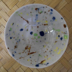 Op de afbeelding: Een witte keramische kom met een gespikkeld ontwerp. De kom heeft kleurrijke spetters van blauw, groen, geel en bruin. De kom is rond en heeft een glanzende afwerking. De kom wordt waarschijnlijk gebruikt voor het serveren van voedsel.