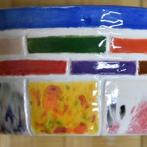 May include: A ceramic planter with a blue rim and a colorful brick-like design. The planter features rectangular blocks in various colors, including red, green, purple, and yellow, creating a vibrant and artistic appearance. The planter is suitable for small plants.