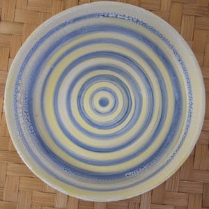 Op de afbeelding: Een ronde keramische plaat met een concentrisch spiraalontwerp. De plaat heeft afwisselende banden van lichtgeel en blauw, waardoor een visueel aantrekkelijk patroon ontstaat. De rand van de plaat heeft een getextureerde, gebroken witte rand.