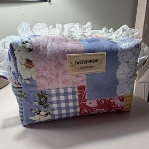 May include: A rectangular fabric pouch featuring a patchwork design of floral, gingham, and solid patterns in shades of blue, pink, red, and yellow. The top edge is trimmed with white lace, and a beige label reads JWLRYBYMIMI - handmade -.