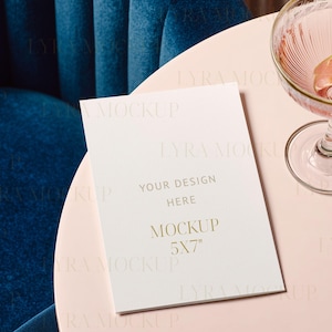 May include: A mockup card with the text "YOUR DESIGN HERE" and "MOCKUP 5X7" in gold lettering. The card is on a pink surface next to a cocktail glass with a citrus slice. A blue velvet chair is in the background.