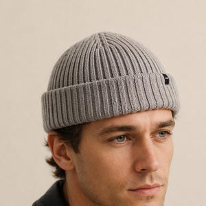 Peut inclure: Un bonnet en tricot côtelé gris clair avec un revers plié et une petite étiquette rectangulaire noire de la marque fixée sur le côté. Le bonnet présente un motif en tricot vertical et une calotte arrondie, offrant une coupe structurée.