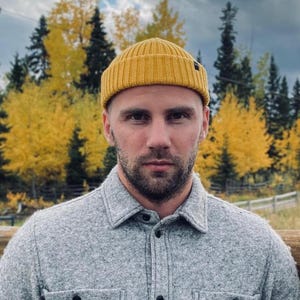 Può includere: Un uomo con la barba che indossa una camicia grigia e un berretto giallo.
