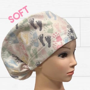 May include: A white scrub cap with a pattern of colorful handprints and footprints. The word "SOFT" is printed in pink across the top. The cap is designed to be worn in a medical setting.