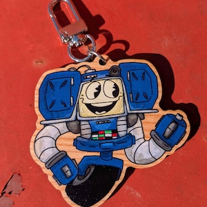 May include: Wooden robot-themed keychain with a silver clasp. The robot has a blue and yellow color scheme with a smiling face. The arms and legs are white and gray. The keychain is on a red surface.