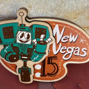 May include: Wooden laser-cut sign featuring a retro robot holding a drink, with the text "New Vegas 15" in a stylized design. The robot is teal and brown, set against an orange and white background. The sign is made of light-coloured wood.