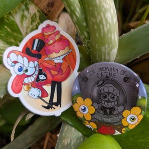 May include: Two stickers and a button featuring cartoon characters. One sticker shows a red character with a top hat and a cake. The button reads "In Memory of Caine" with a tombstone design and the years 1996-2026.