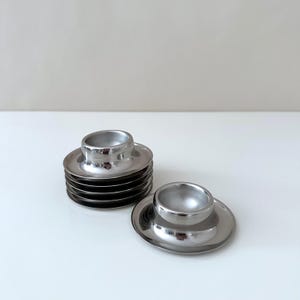 Peut inclure: Ensemble de coquetiers argentés brillants. Chaque coquetier est composé d'un petit bol arrondi posé sur une base circulaire. Une pile de coquetiers est visible, ainsi que deux coquetiers individuels. Les surfaces réfléchissantes créent des reflets.