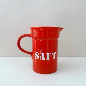 Könnte beinhalten: Glänzender roter Keramikkrug mit Henkel und Ausguss. Das Wort "SAFT" ist weiß auf der Vorderseite aufgedruckt. Der Krug steht auf einer weißen Oberfläche.
