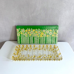 May include: A rectangular, clear glass tray with rounded amber-colored beads around the edges and interior. A green box with a floral pattern in yellow, red, and green sits behind the tray. The tray and box are on a white surface.