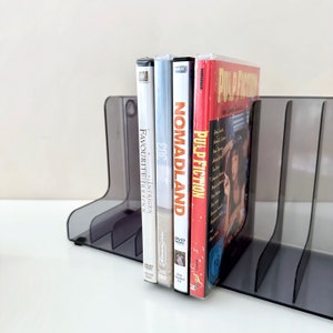 May include: A black plastic DVD holder with four slots, holding four DVD cases. The DVD cases are all different colours and have different titles. The titles are "Pulp Fiction", "Nomadland", "The Intriguing Irrsinn", and "Favourite".
