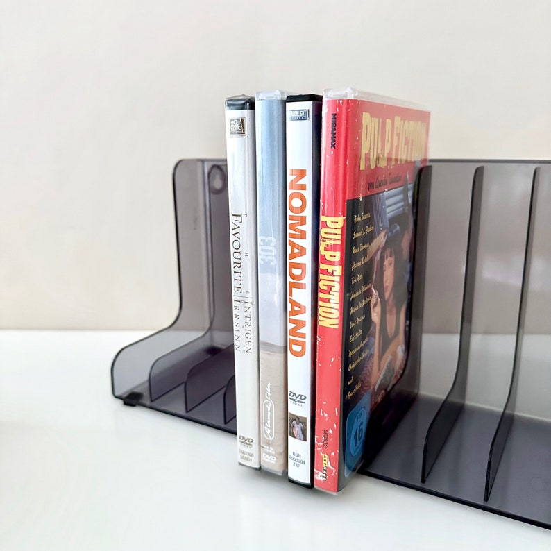 May include: A black plastic DVD holder with four slots, holding four DVD cases. The DVD cases are all different colours and have different titles. The titles are "Pulp Fiction", "Nomadland", "The Intriguing Irrsinn", and "Favourite".